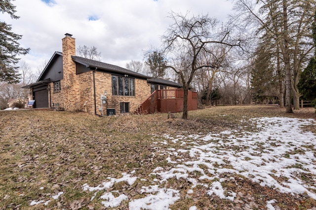 view of snowy exterior featuring a garage, brick siding, a chimney, and a wooden deck