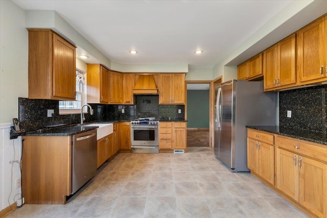 kitchen featuring premium range hood, a sink, appliances with stainless steel finishes, dark stone counters, and brown cabinetry