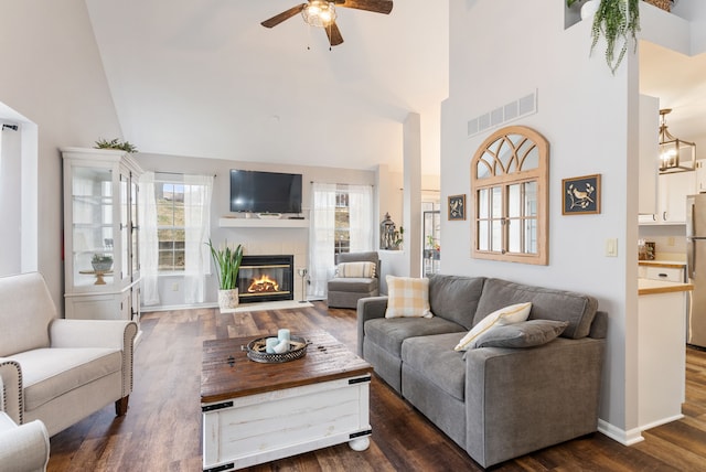 living area featuring ceiling fan, a towering ceiling, dark wood finished floors, visible vents, and a glass covered fireplace