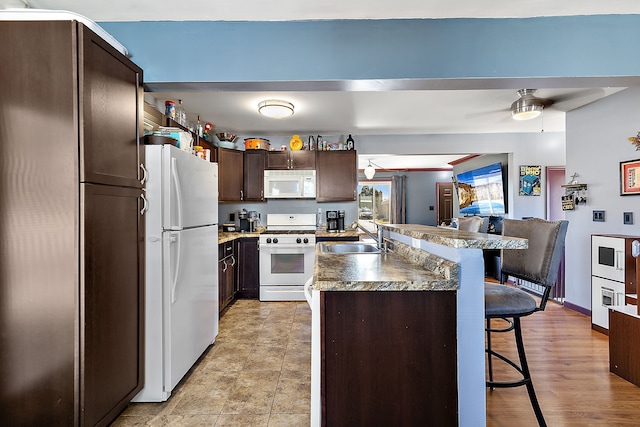 kitchen with a breakfast bar, a sink, dark brown cabinetry, white appliances, and baseboards