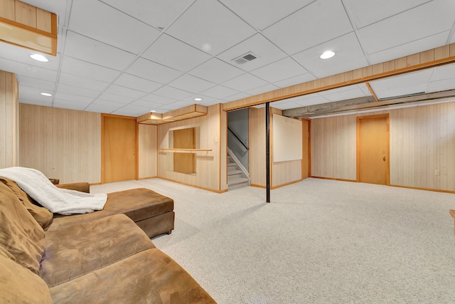 carpeted living area featuring baseboards, visible vents, a drop ceiling, stairs, and wood walls