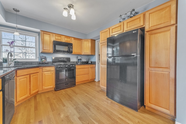 kitchen with dark stone counters, a sink, hanging light fixtures, black appliances, and light wood-type flooring