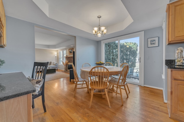 dining space featuring baseboards, a tray ceiling, light wood-style floors, a glass covered fireplace, and a notable chandelier