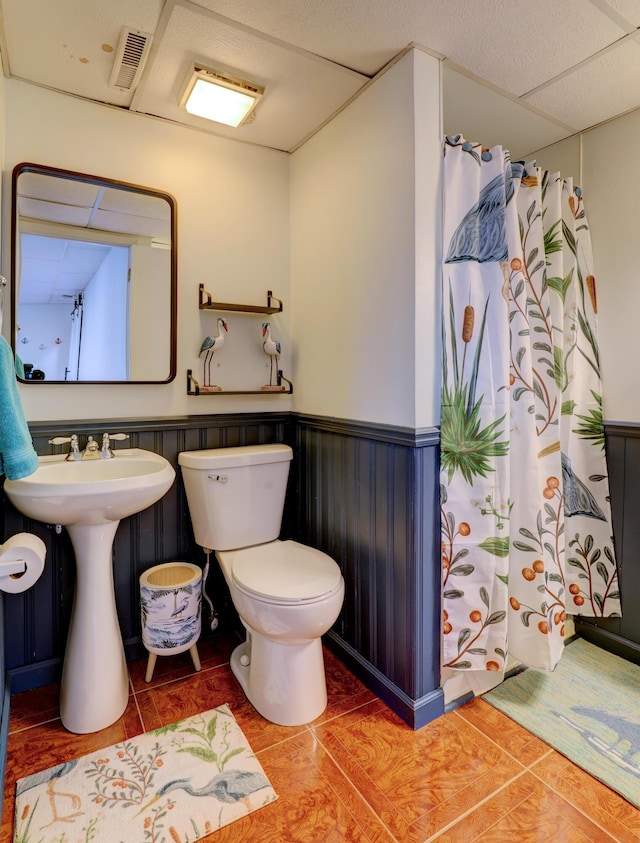 bathroom with a wainscoted wall, curtained shower, visible vents, toilet, and a drop ceiling