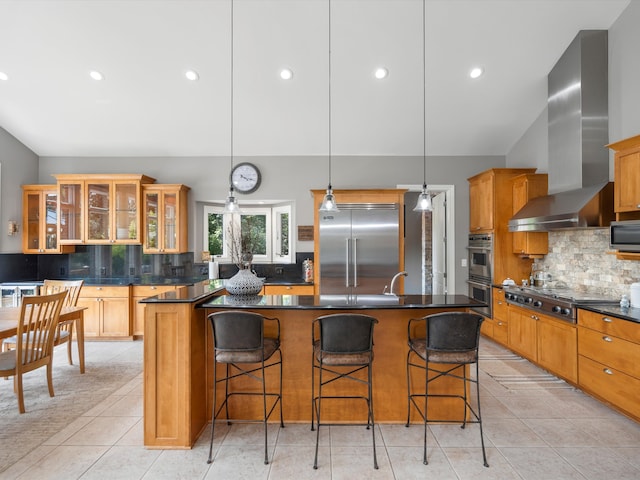 kitchen featuring appliances with stainless steel finishes, dark countertops, wall chimney range hood, and an island with sink