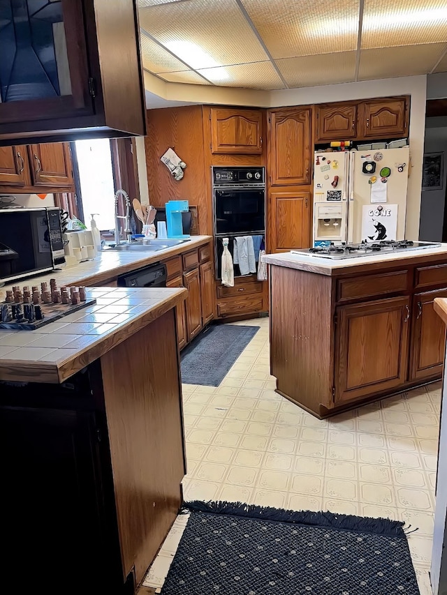 kitchen featuring light floors, a sink, tile counters, brown cabinets, and black appliances