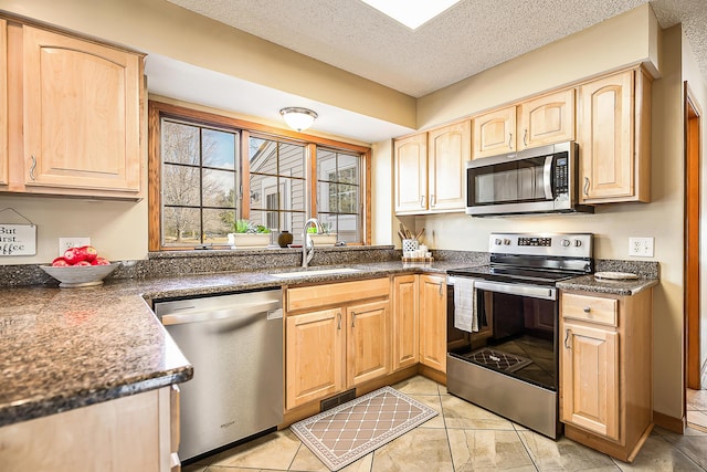 kitchen with a textured ceiling, a sink, appliances with stainless steel finishes, light brown cabinetry, and dark countertops