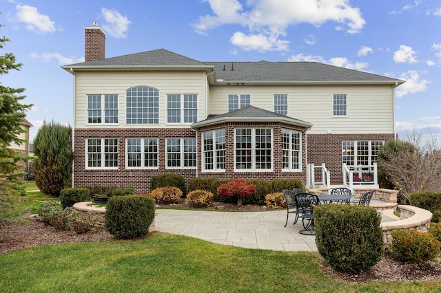 back of house featuring roof with shingles, brick siding, a chimney, a lawn, and a patio area