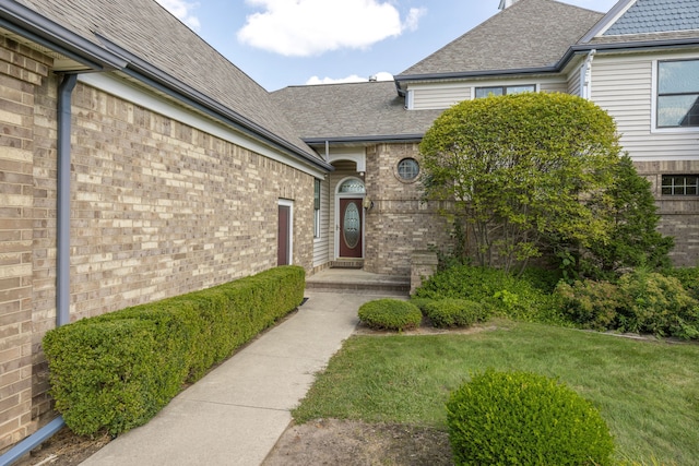 doorway to property featuring a shingled roof, brick siding, and a lawn