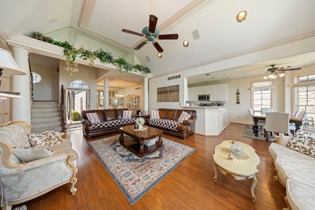 living room featuring high vaulted ceiling, ceiling fan with notable chandelier, wood finished floors, stairs, and decorative columns