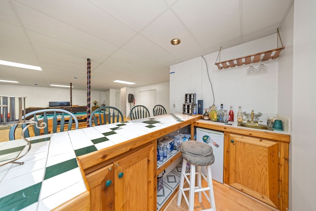 kitchen with tile counters, a sink, light wood-type flooring, a drop ceiling, and dishwasher