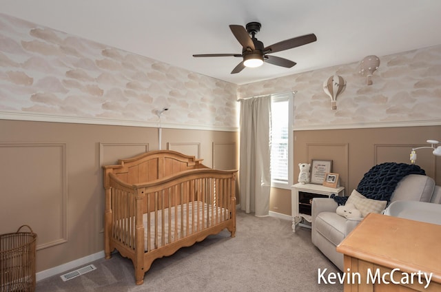 carpeted bedroom featuring visible vents, a decorative wall, wainscoting, ceiling fan, and a crib