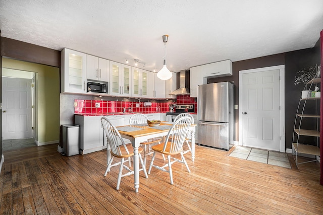 kitchen featuring stainless steel appliances, white cabinets, wall chimney range hood, light wood finished floors, and tasteful backsplash