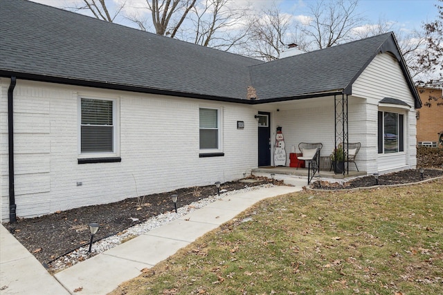 view of front facade with roof with shingles, covered porch, a chimney, a front lawn, and brick siding