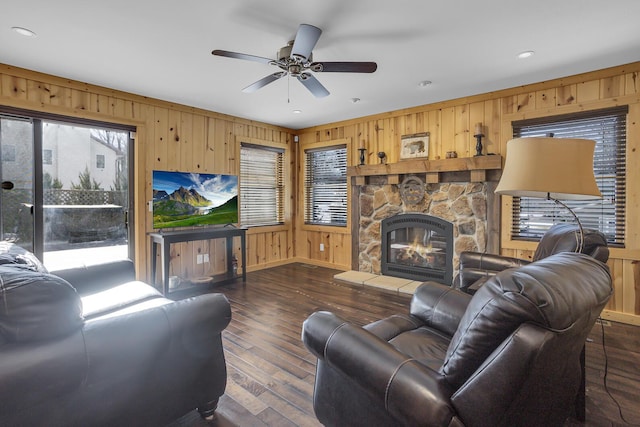 living room featuring wooden walls, wood-type flooring, ceiling fan, and a stone fireplace