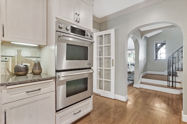 kitchen featuring light wood finished floors, tasteful backsplash, dark stone countertops, crown molding, and stainless steel double oven