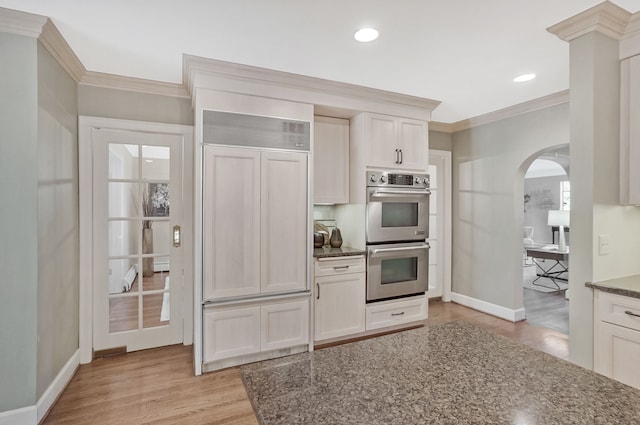 kitchen featuring arched walkways, double oven, paneled built in fridge, ornamental molding, and light wood-type flooring