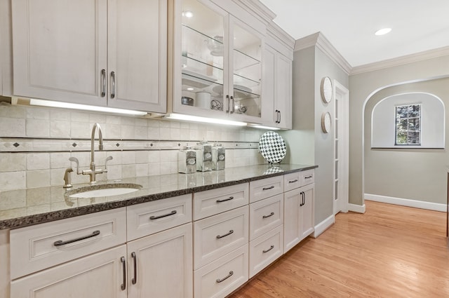 kitchen with dark stone counters, glass insert cabinets, crown molding, light wood-style floors, and a sink