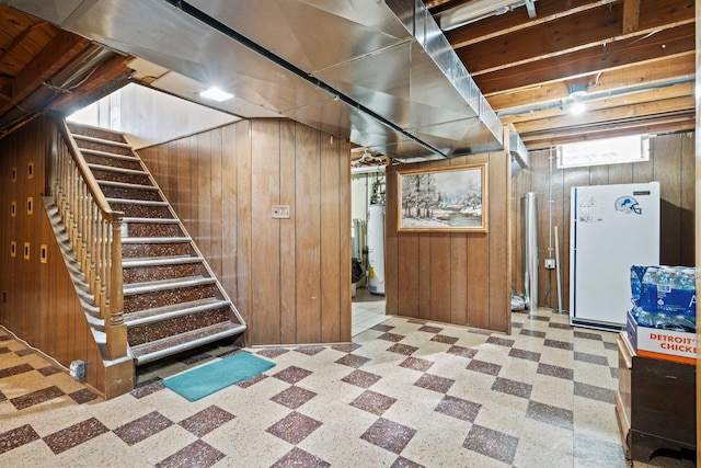 basement featuring tile patterned floors, water heater, freestanding refrigerator, wood walls, and stairs