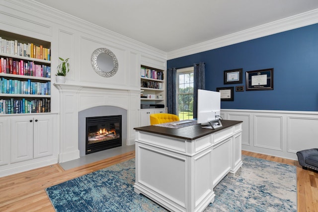 office area with built in shelves, crown molding, light wood finished floors, a fireplace with flush hearth, and wainscoting