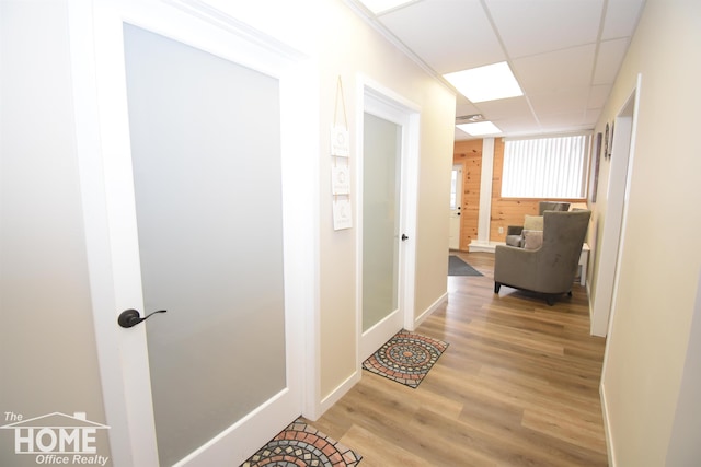 hall featuring light wood-style floors, wooden walls, baseboards, and a drop ceiling