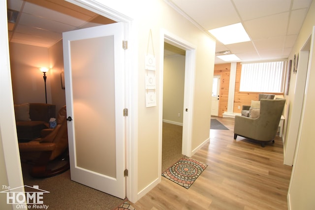 corridor with light wood-type flooring, a paneled ceiling, wood walls, and baseboards