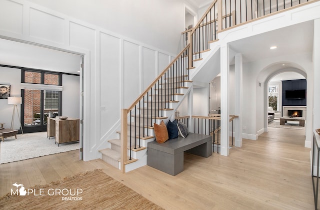 stairway featuring a warm lit fireplace, arched walkways, wood finished floors, a high ceiling, and a decorative wall