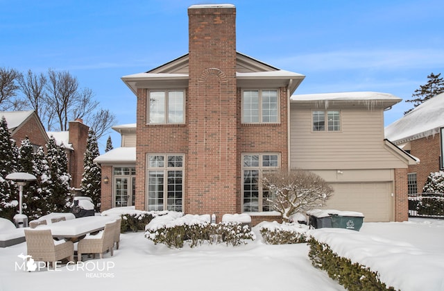 snow covered rear of property featuring an attached garage, outdoor dining space, a chimney, and brick siding