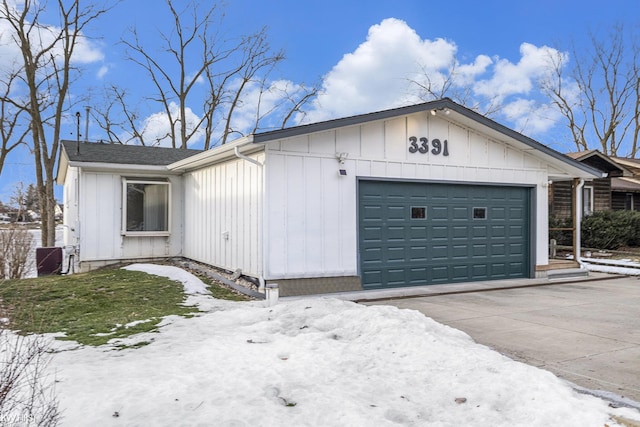 view of snowy exterior with concrete driveway, board and batten siding, an attached garage, and roof with shingles