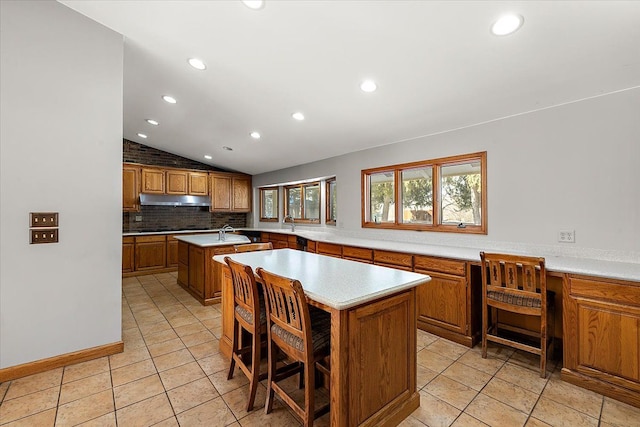 kitchen with under cabinet range hood, vaulted ceiling, light countertops, brown cabinetry, and a center island with sink