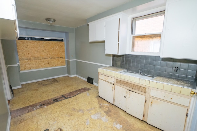 kitchen with tile countertops, backsplash, white cabinetry, a sink, and baseboards