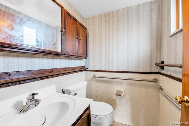 half bath featuring toilet, a wainscoted wall, tile walls, vanity, and wallpapered walls