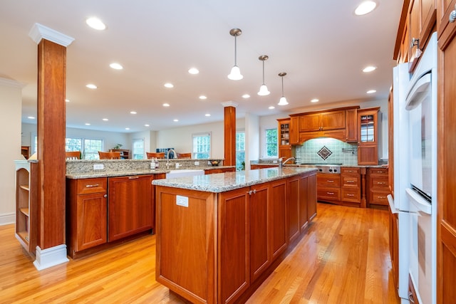 kitchen with brown cabinetry, an island with sink, ornate columns, and light wood finished floors