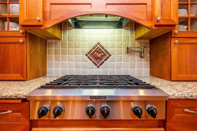 details with stainless steel gas cooktop, tasteful backsplash, brown cabinetry, and glass insert cabinets