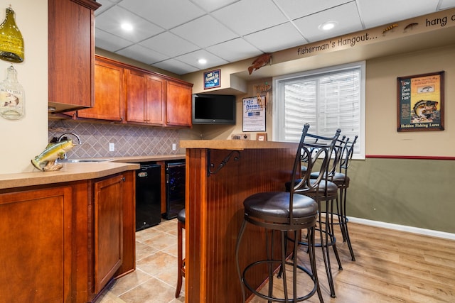 kitchen featuring beverage cooler, a sink, a drop ceiling, a kitchen breakfast bar, and decorative backsplash