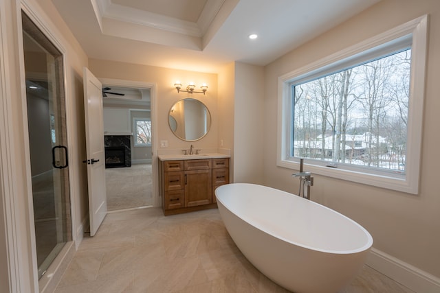 bathroom with crown molding, a premium fireplace, a raised ceiling, a soaking tub, and vanity