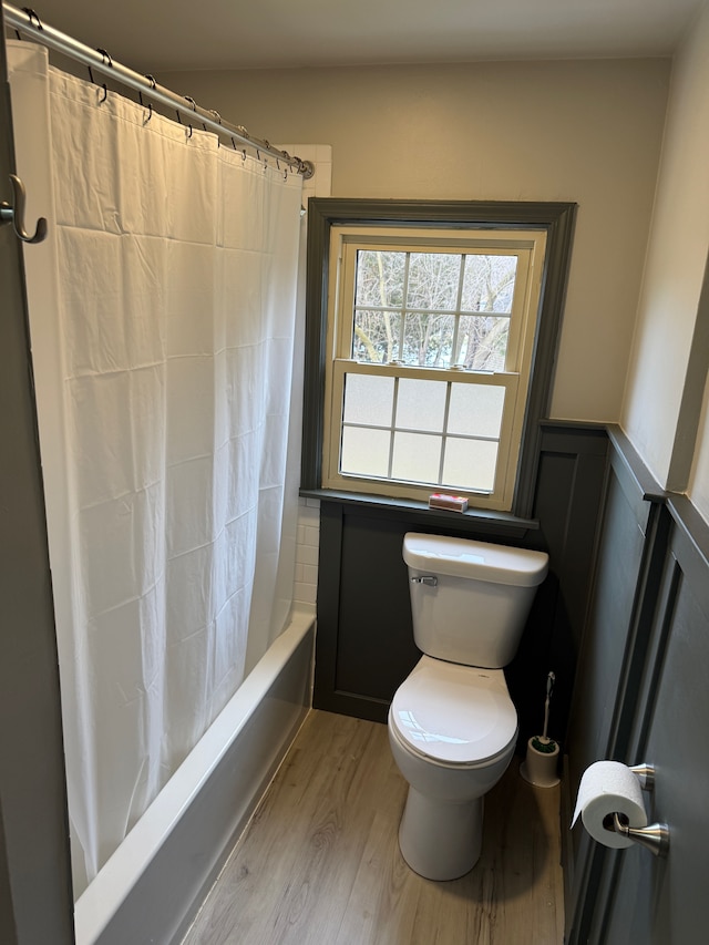 bathroom with a wainscoted wall, shower / bath combination with curtain, a decorative wall, toilet, and wood finished floors