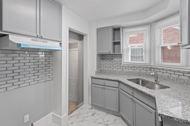 kitchen with light stone counters, gray cabinetry, under cabinet range hood, a sink, and marble finish floor