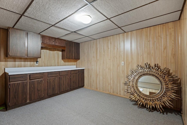 kitchen with light carpet, dark brown cabinetry, light countertops, a paneled ceiling, and wood walls