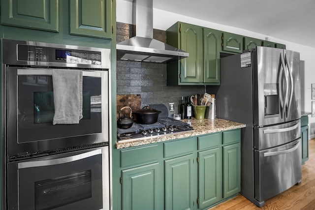 kitchen with light wood finished floors, stainless steel appliances, wall chimney range hood, green cabinets, and backsplash