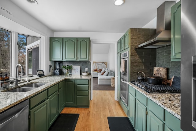 kitchen with light wood finished floors, appliances with stainless steel finishes, a sink, wall chimney range hood, and green cabinetry