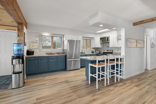 kitchen with blue cabinets, a peninsula, appliances with stainless steel finishes, light wood-type flooring, and beamed ceiling