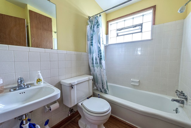 bathroom featuring tile walls, wainscoting, toilet, shower / bath combo with shower curtain, and a sink