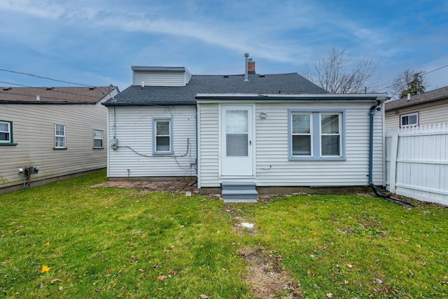 rear view of house featuring a chimney, a shingled roof, a lawn, entry steps, and fence