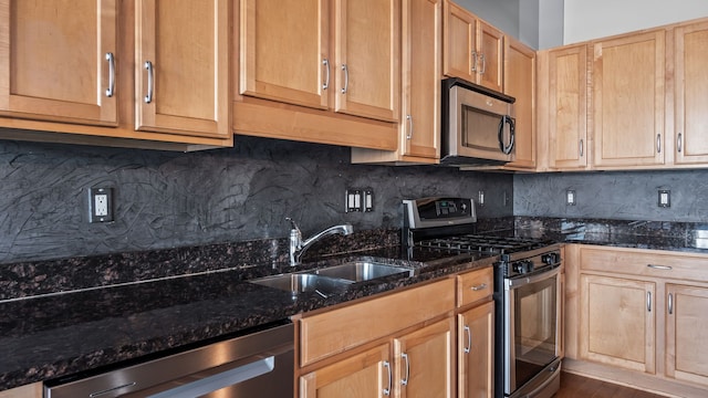 kitchen with stainless steel appliances, a sink, backsplash, dark stone counters, and light brown cabinetry