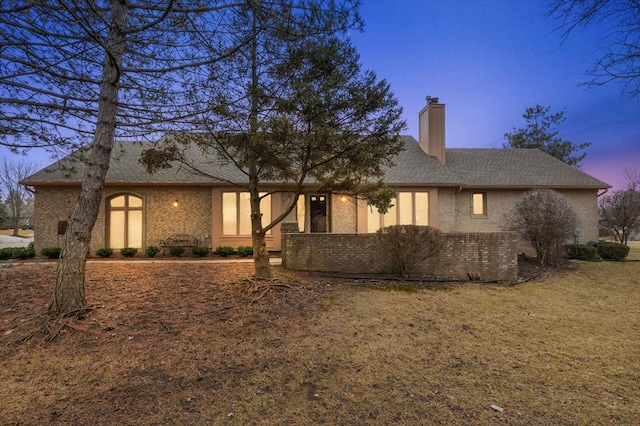 back of property at dusk featuring brick siding, a chimney, and roof with shingles