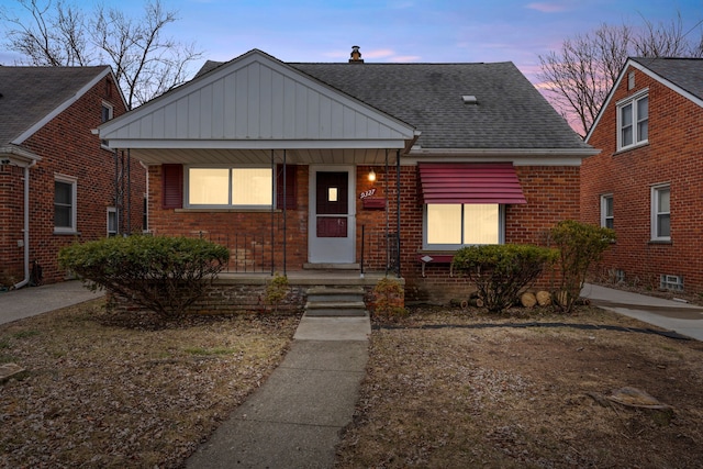 bungalow featuring covered porch, brick siding, and roof with shingles