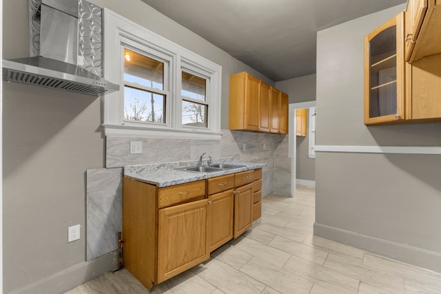 kitchen featuring tasteful backsplash, glass insert cabinets, a sink, wall chimney range hood, and baseboards