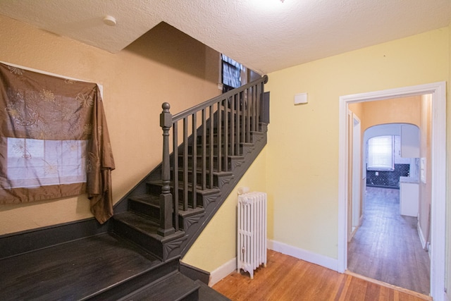 stairs with arched walkways, a textured ceiling, wood finished floors, baseboards, and radiator heating unit