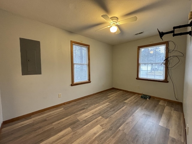 unfurnished room with a textured ceiling, wood finished floors, visible vents, baseboards, and electric panel